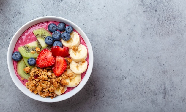 Close-up de tigelas de suco de açaí de verão com morangos, banana, mirtilos, kiwis e granola em fundo cinza de concreto. tigela de café da manhã com frutas e cereais, vista de cima, espaço para texto