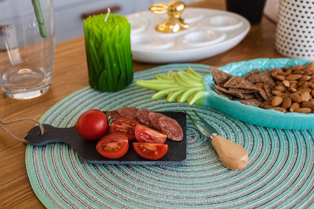 Close-up de tomate cereja fresco com linguiça fatiada a bordo, travessa de amêndoas e biscoitos no balcão de madeira com vela em forma e travessa de ovos com coelhinho da Páscoa por cima.