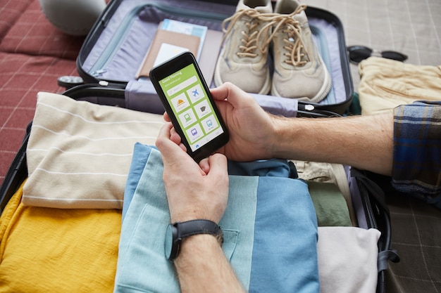 Close-up de turista usando celular para viajar, sentado no sofá com uma mala com roupas prontas