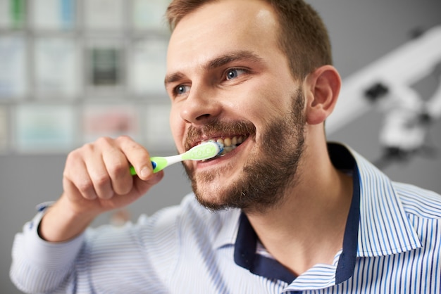 Close-up de um cara com uma barba na camisa limpa os dentes no consultório odontológico