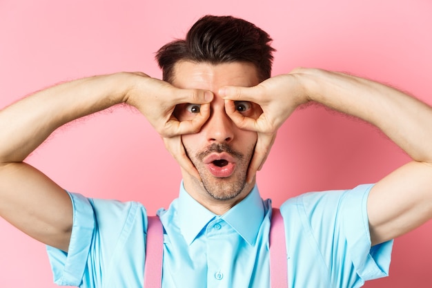 Close-up de um cara engraçado caucasiano com bigode e cerdas, de pé em suspensórios e fazendo máscara de super-herói com as mãos sobre os olhos, em pé sobre um fundo rosa.