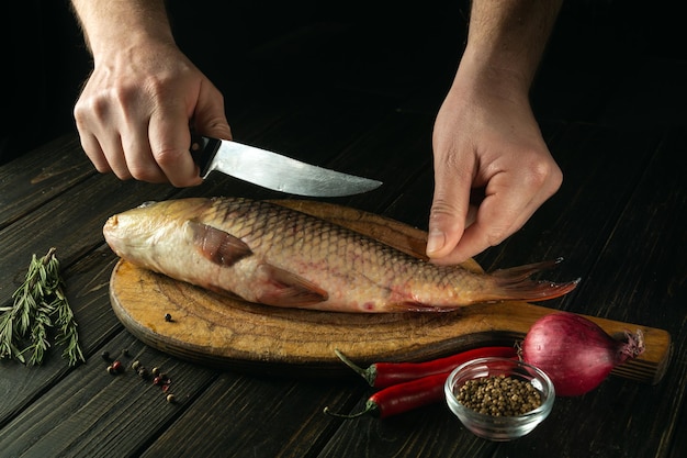 Close-up de um cozinheiro mãos com uma faca na mesa da cozinha antes de cortar carpa crua Cozinhar um prato de peixe na mesa da cozinha com especiarias e pimenta