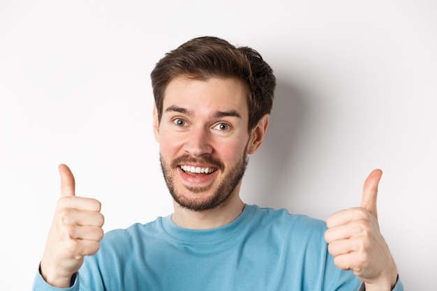 Close-up de um homem alegre dizer sim, mostrando os polegares em aprovação, elogie o bom trabalho, sorrindo com aprovação, de pé sobre um fundo branco.