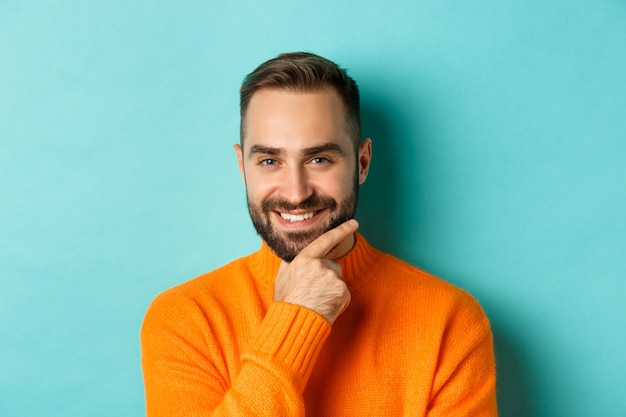 Close-up de um homem bonito e confiante com barba sorrindo, olhando pensativo para a câmera, tem um plano, em pé sobre um fundo azul claro.