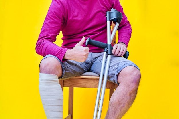 Foto close-up de um homem com muletas, jeans e camisa roxa, sentado em uma cadeira com o polegar para cima.