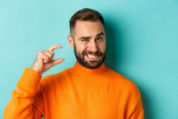 Close-up de um homem estranho aparecendo pequeno ou pequeno com os dedos, em pé descontente com o fundo azul claro.