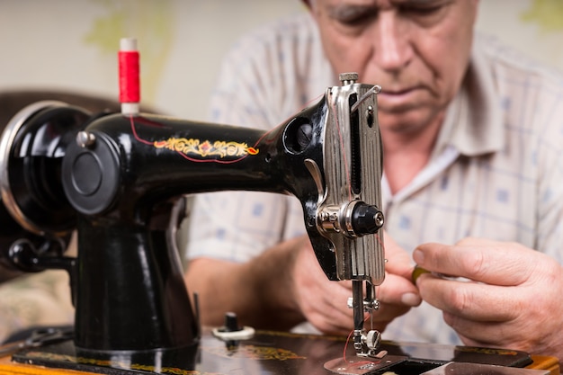 Close-up de um homem sênior enfiando a linha na máquina de costura manual à moda antiga com fio vermelho