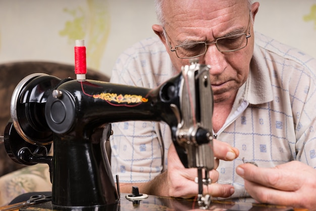 Close-up de um homem sênior enfiando a linha na máquina de costura manual à moda antiga com fio vermelho