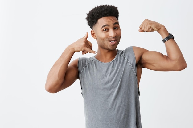 Close-up de um jovem alegre, de pele escura, com corte de cabelo afro, uma camisa cinza mostrando os músculos, fazendo sinal de telefone com a mão