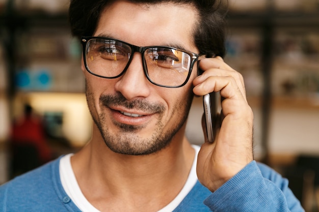 Close-up de um jovem bonito usando óculos na biblioteca, falando no celular