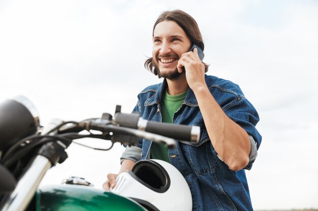 Close-up de um jovem bonito vestindo roupa casual sentado em uma motocicleta na praia, falando ao telefone celular