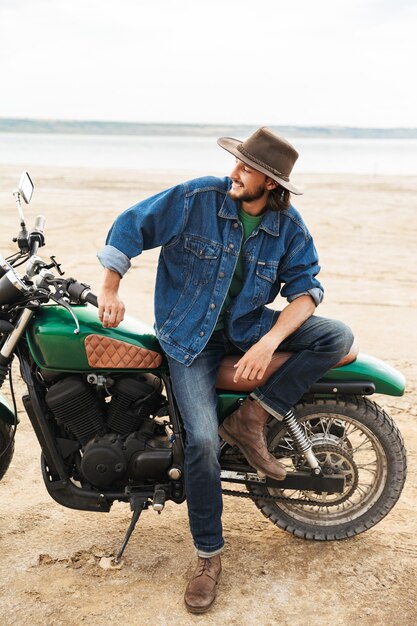 Close-up de um jovem bonito vestindo roupa casual sentado em uma motocicleta na praia