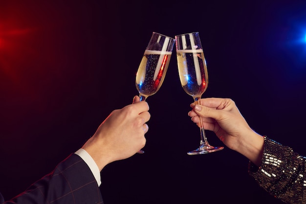 Close-up de um jovem casal tilintando taças de champanhe iluminadas por luzes de festa contra um fundo preto filmado com flash