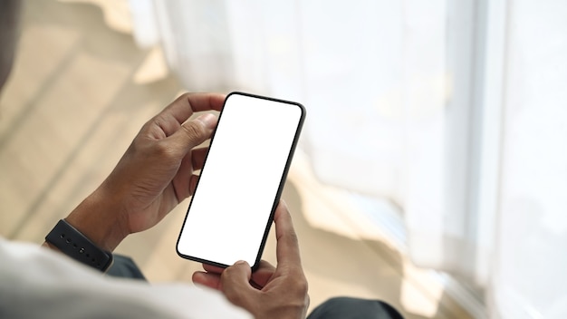 Close-up de um jovem sentado perto da janela segurando uma simulação de telefone inteligente com tela branca