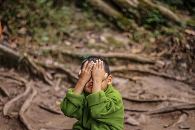 Foto close-up de um menino com as mãos cobrindo os olhos sentado em terra