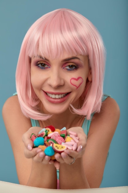 Close-up de um modelo feminino paquerador em fundo azul usando uma peruca rosa segurando diferentes tipos de doces Mulher muito glamourosa cabelo rosa charme doces estilo de vida prazer açúcar