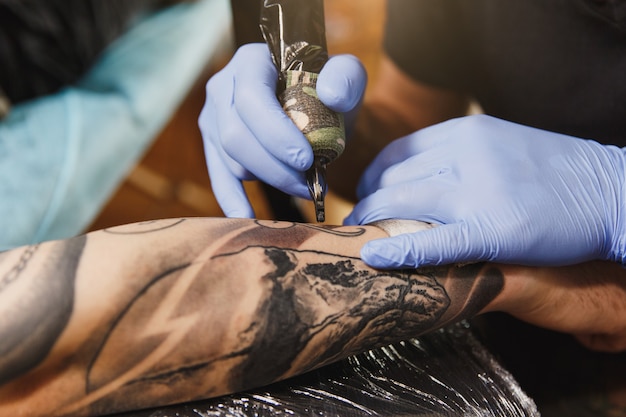 Close-up de um tatuador profissional fazendo uma tatuagem no braço de um jovem por uma máquina com tinta preta