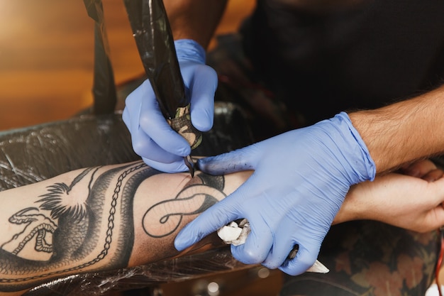Close-up de um tatuador profissional fazendo uma tatuagem no braço de um jovem por uma máquina com tinta preta