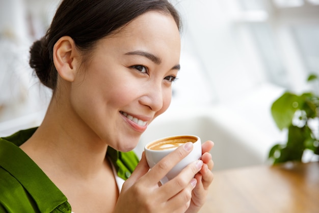 Close-up de uma adorável jovem asiática tomando café enquanto está sentado em uma cafeteria dentro de casa