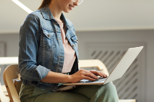 Close-up de uma aluna sorridente com jaqueta jeans, sentada na cadeira e digitando no laptop na universidade