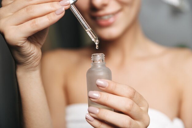 Foto close-up de uma bela jovem enrolada em uma toalha aplicando óleo cosmético de um frasco no rosto