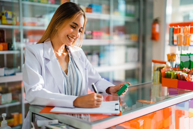 Close-up de uma farmacêutica asiática trabalhando escrevendo uma lista de medicamentos com caneta e prancheta na farmácia