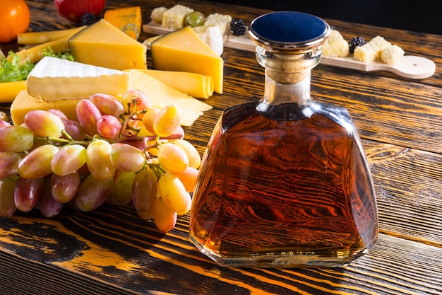 Close-up de uma garrafa de álcool em uma mesa de madeira rústica com uma variedade de queijos e uvas com iluminação lateral brilhante