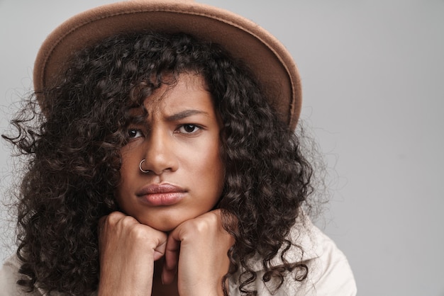 Close-up de uma jovem africana atraente, chateada, com cabelo encaracolado, vestindo um casaco de outono e um chapéu