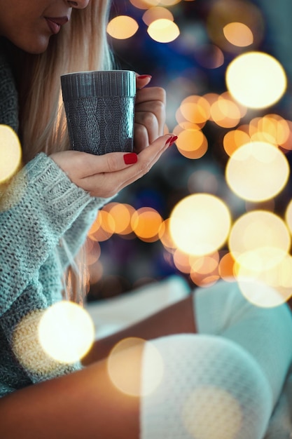 Close-up de uma jovem bonita está sentada na cama e segurando uma xícara de chá ou café, rodeada de luzes de Natal.