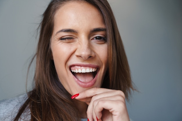 Foto close-up de uma jovem brincalhona linda de pé isolada sobre uma parede cinza, piscando