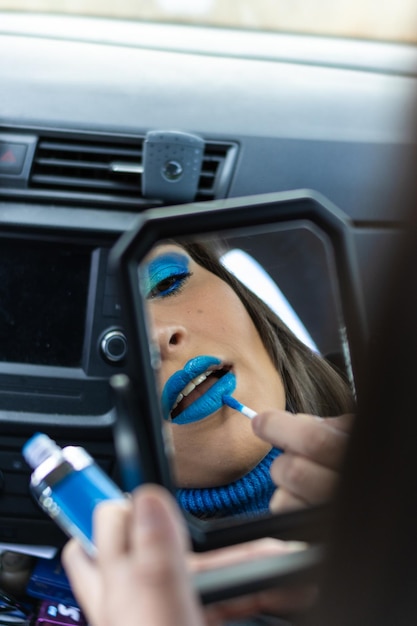 Close-up de uma jovem fazendo sua maquiagem dentro de um carro com tons de azul, olhando para um espelho