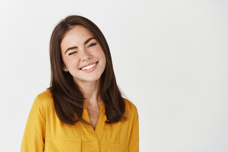 Foto close-up de uma jovem feliz piscando para a câmera, sorrindo, alegre, em pé sobre uma parede branca