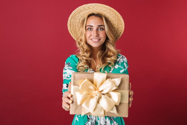 Close-up de uma jovem loira atraente e feliz usando um vestido de verão e chapéu de palha em pé, isolado sobre uma parede rosa, mostrando a caixa de presente