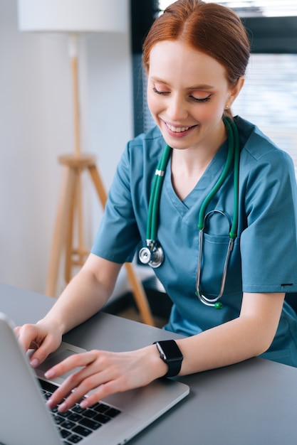 Close-up de uma jovem médica muito sorridente em uniforme médico verde azul, sentada na mesa com o laptop no fundo da janela