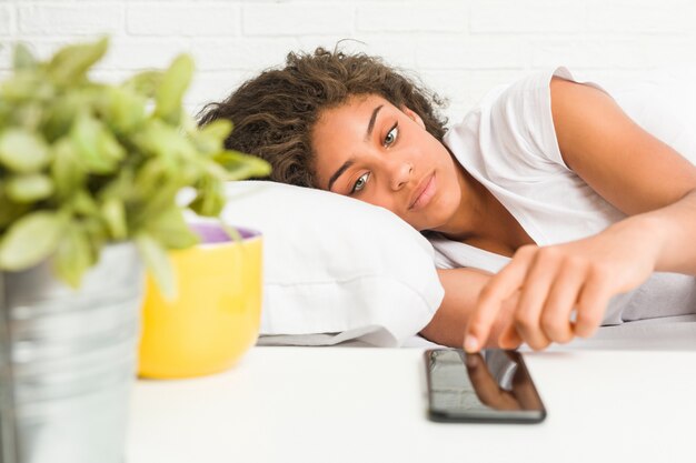 Close-up de uma jovem mulher afro-americana, deitada na cama, olhando um telefone