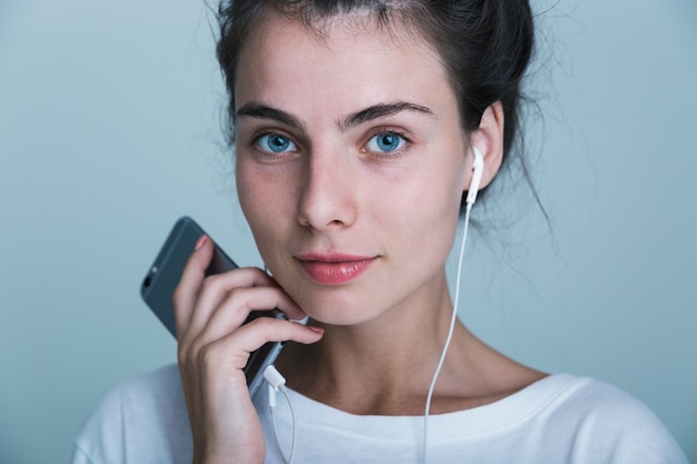 Close-up de uma linda jovem casual isolada sobre um fundo azul, ouvindo música com fones de ouvido e telefone celular