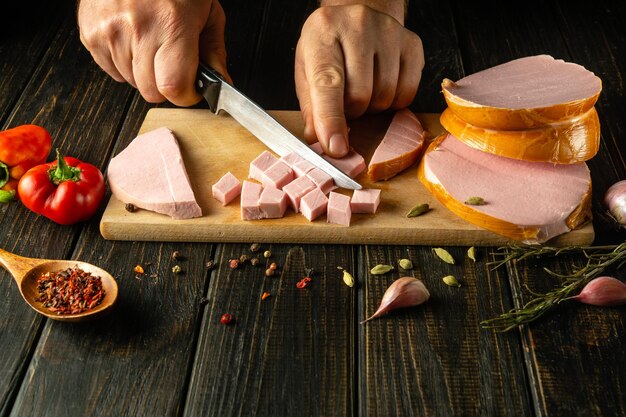 Close-up de uma mão de cozinheiro cortando salsicha escaldada em pedaços pequenos com uma faca para preparar um prato de carne