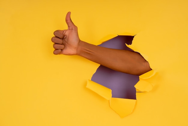 Close-up de uma mão fazendo o polegar para cima gesto com a mão através da parede de papel rasgado.