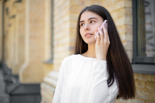 Close-up de uma menina morena pensativa em branco com cabelos longos, falando em um telefone celular ao caminhar pela rua. lugar para o seu design