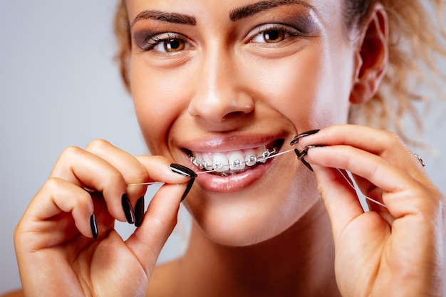 Foto close-up de uma menina sorridente com aparelho limpando os dentes com fio dental. olhando para a câmera.