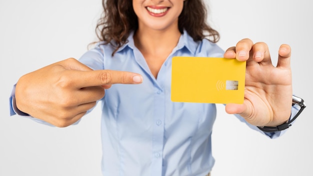 Foto close-up de uma mulher alegre em uma blusa azul claro apontando para um cartão de crédito amarelo