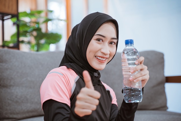 Close-up de uma mulher asiática em um véu sportswear sorri enquanto segura uma garrafa de bebida com os polegares para cima após se exercitar em casa