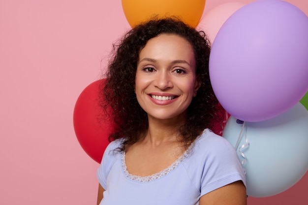 Close-up de uma mulher atraente, feliz, alegre, mestiça, sorrindo com um lindo sorriso cheio de dentes, olhando para a câmera, posando contra balões de ar inflados em um fundo rosa colorido com espaço de cópia