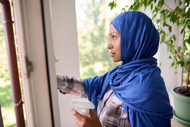 Close-up de uma mulher bonita jovem árabe muçulmana confiante com a cabeça coberta por hijab azul lavando janelas, removendo manchas e limpando com pano durante a limpeza de primavera em casa. copie o espaço para anúncio