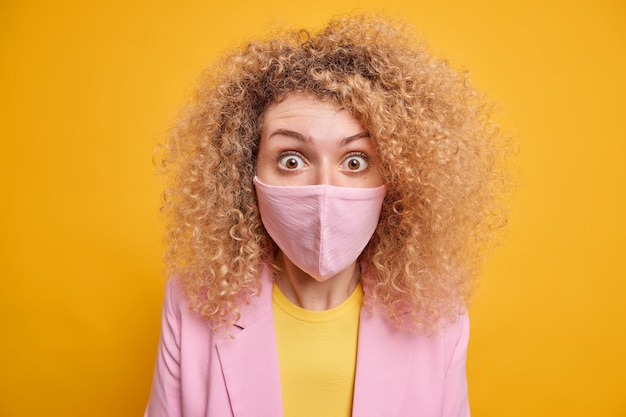 Close up de uma mulher de cabelo encaracolado usando máscara protetora chocada com as restrições de bloqueio e quarentena estende o risco de disseminação de infecção vestida com roupas formais isoladas sobre a parede amarela