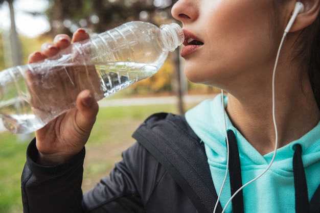 Close-up de uma mulher fitness ouvindo música com fones de ouvido, segurando uma garrafa de água enquanto estava no parque