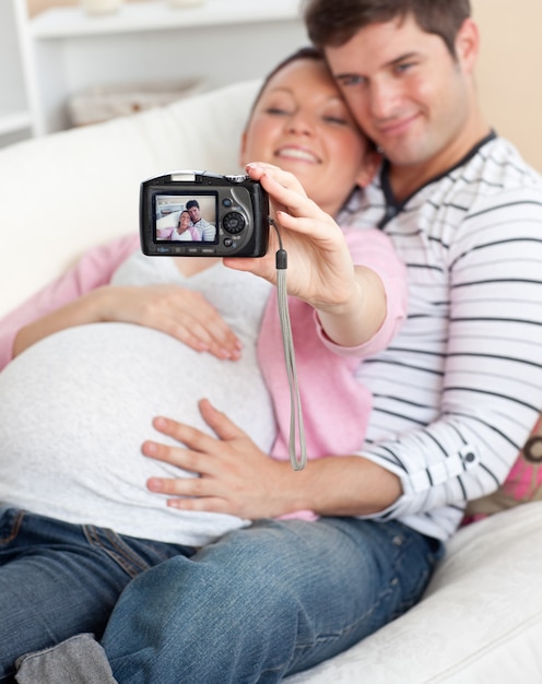 Close up de uma mulher grávida alegre e seu marido tirando fotos de si mesmos em um sofá