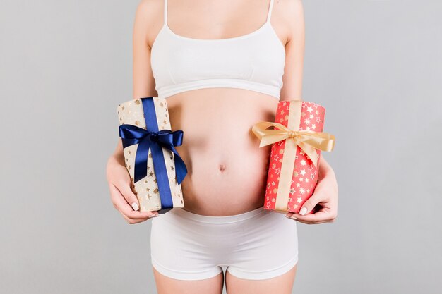 Close-up de uma mulher grávida em cueca branca segurando duas caixas de presente na superfície cinza