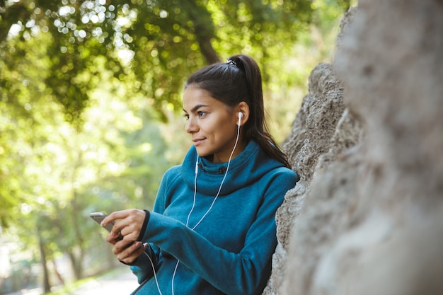 Close-up de uma mulher jovem e atraente fitness vestindo roupas esportivas, fazendo exercícios ao ar livre, ouvindo música com fones de ouvido