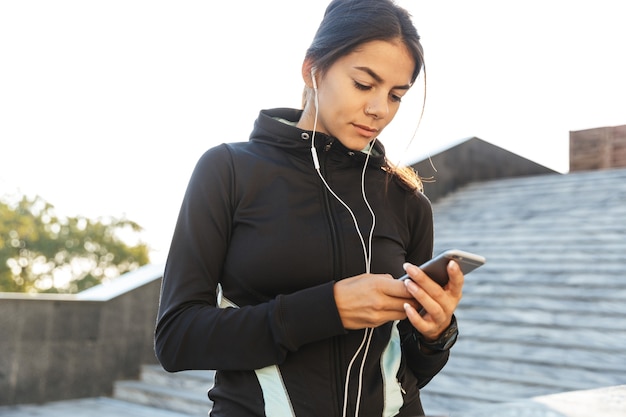 Close-up de uma mulher jovem e atraente fitness vestindo roupas esportivas, fazendo exercícios ao ar livre, usando telefone celular